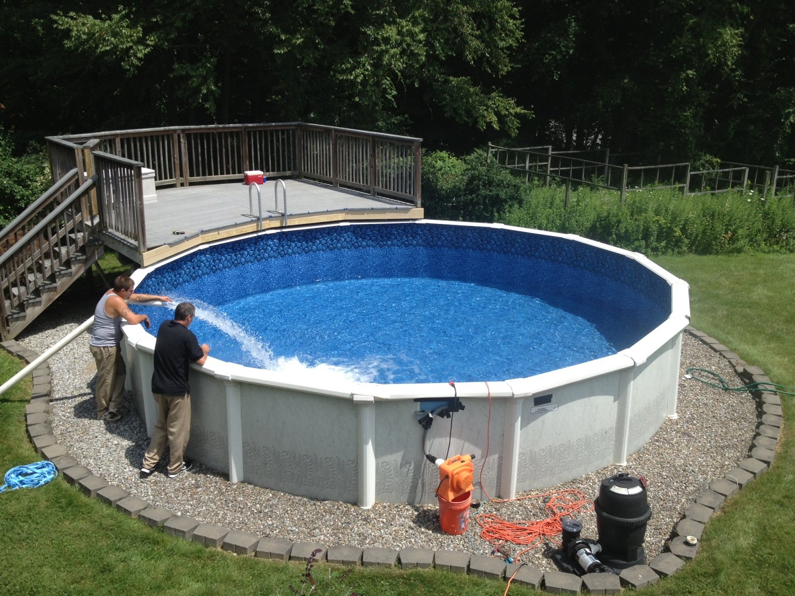 Two installers filling a newly built above-ground swimming pool with an attached wooden deck in a backyard